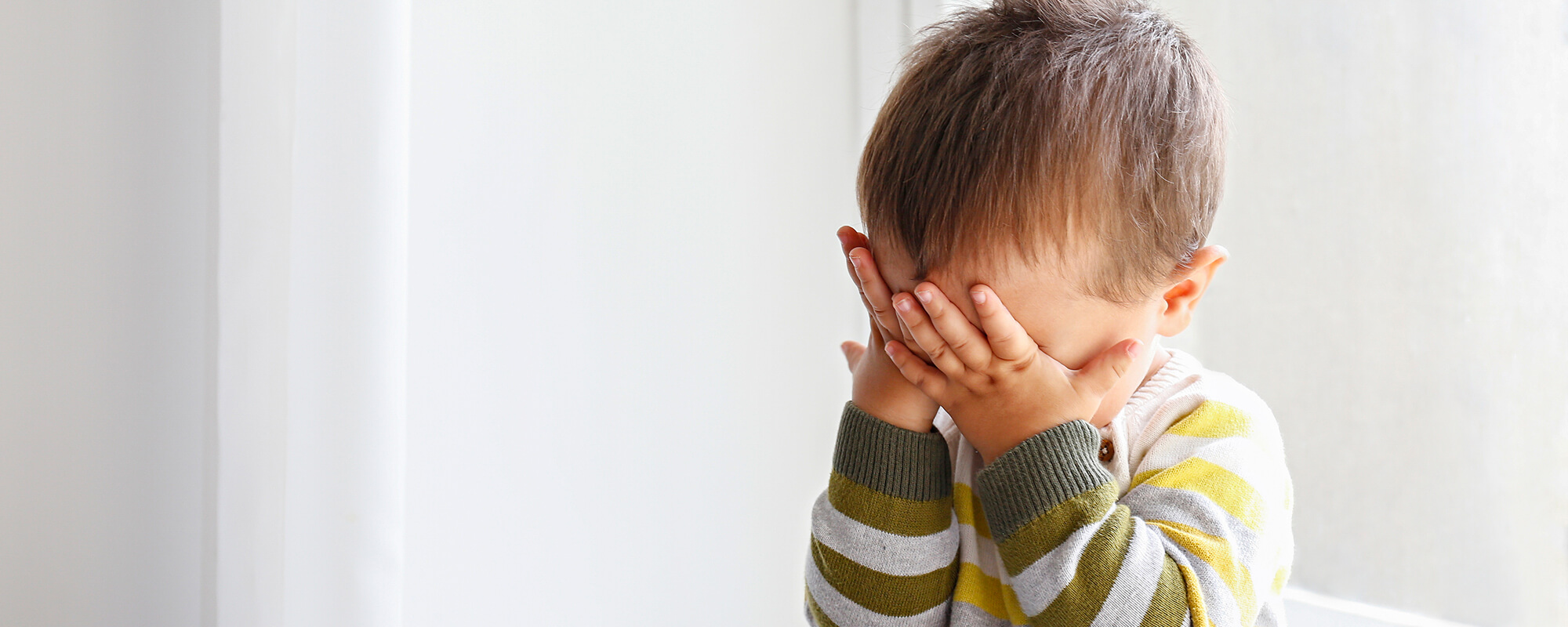 A toddler sits with their hands covering their face.